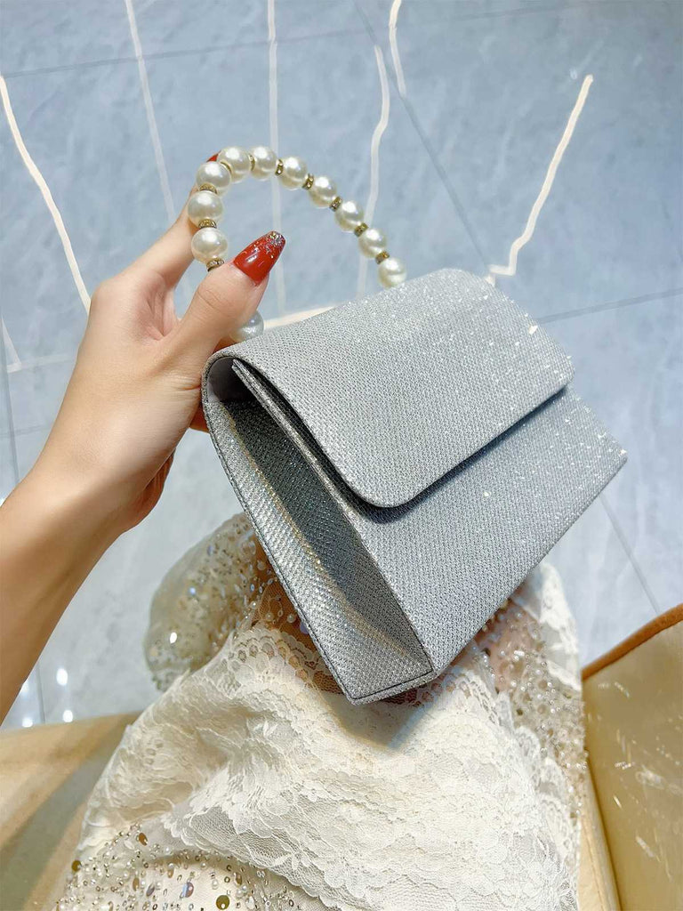 Silberne Clutch, gehalten von einer Hand mit Perlenhenkel, vor einer strukturierten Oberfläche